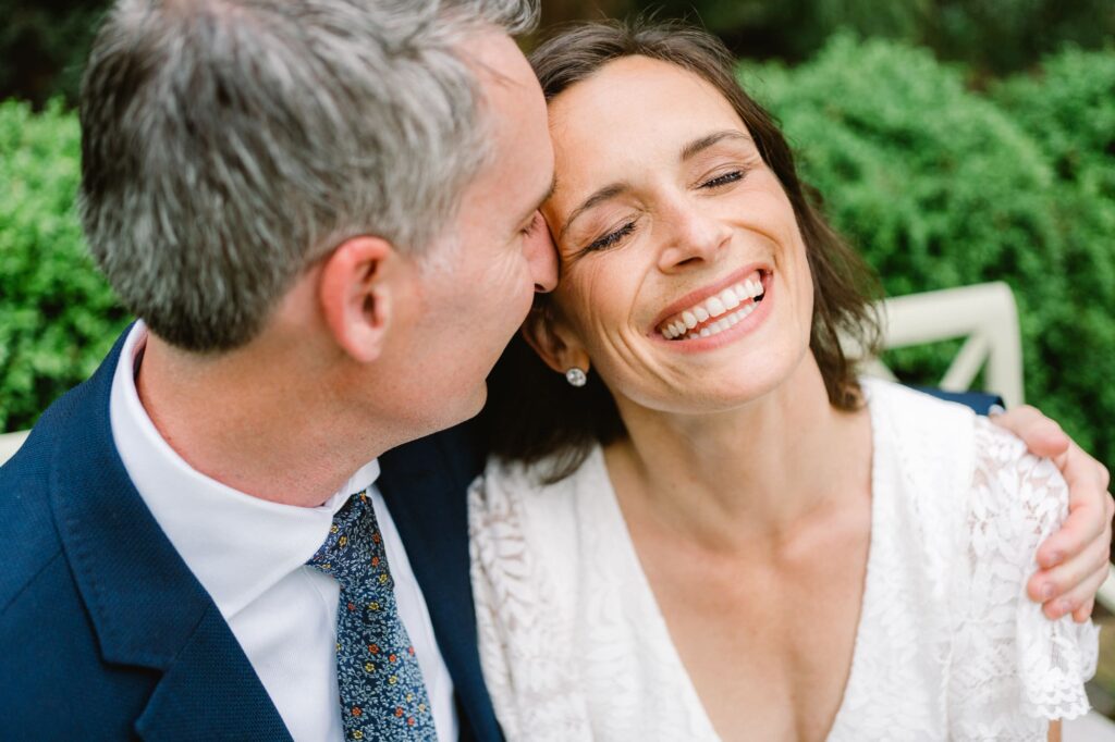 Bride smiles as groom nuzzles her in gardens at Waterperry Farm wedding